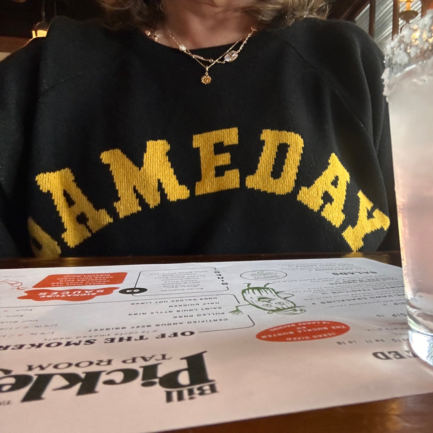 Black sweatshirt with yellow text on a table next to a glass container and a box labeled 'Bill Pickles Tap Room'.