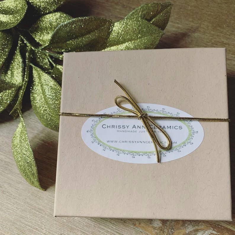 Gift box with a bow and 'Chrissy Anne Ceramics' label on a wooden surface with green leaves.