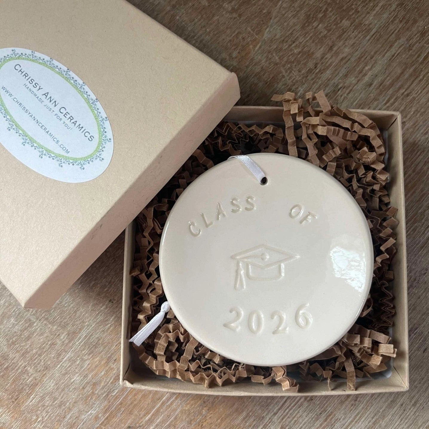 Ceramic disc with 'Class of 2026' engraving in a box with crinkled paper lining.