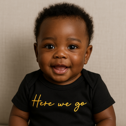Baby wearing a black onesie with 'Here we go' in yellow text, sitting on a beige surface.