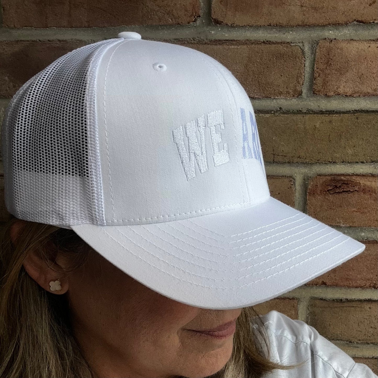 A white trucker-style baseball cap with mesh back panels and embroidered text on the front, worn by a person in front of a brick wall.