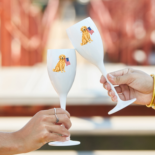 Two white wine glasses with dog illustrations and American flags held by hands against a blurred outdoor background.