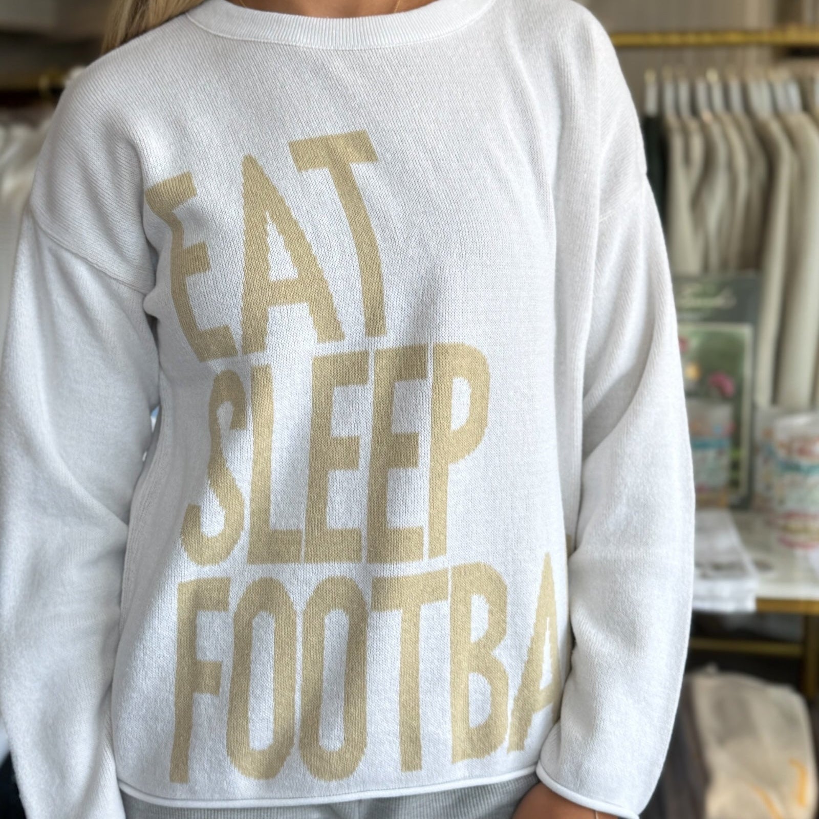 White sweatshirt with 'EAT SLEEP FOOTBALL' text in gold worn by a person indoors.