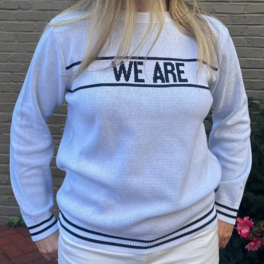 Woman wearing a white sweater with 'WE ARE' text, standing against a brick wall.