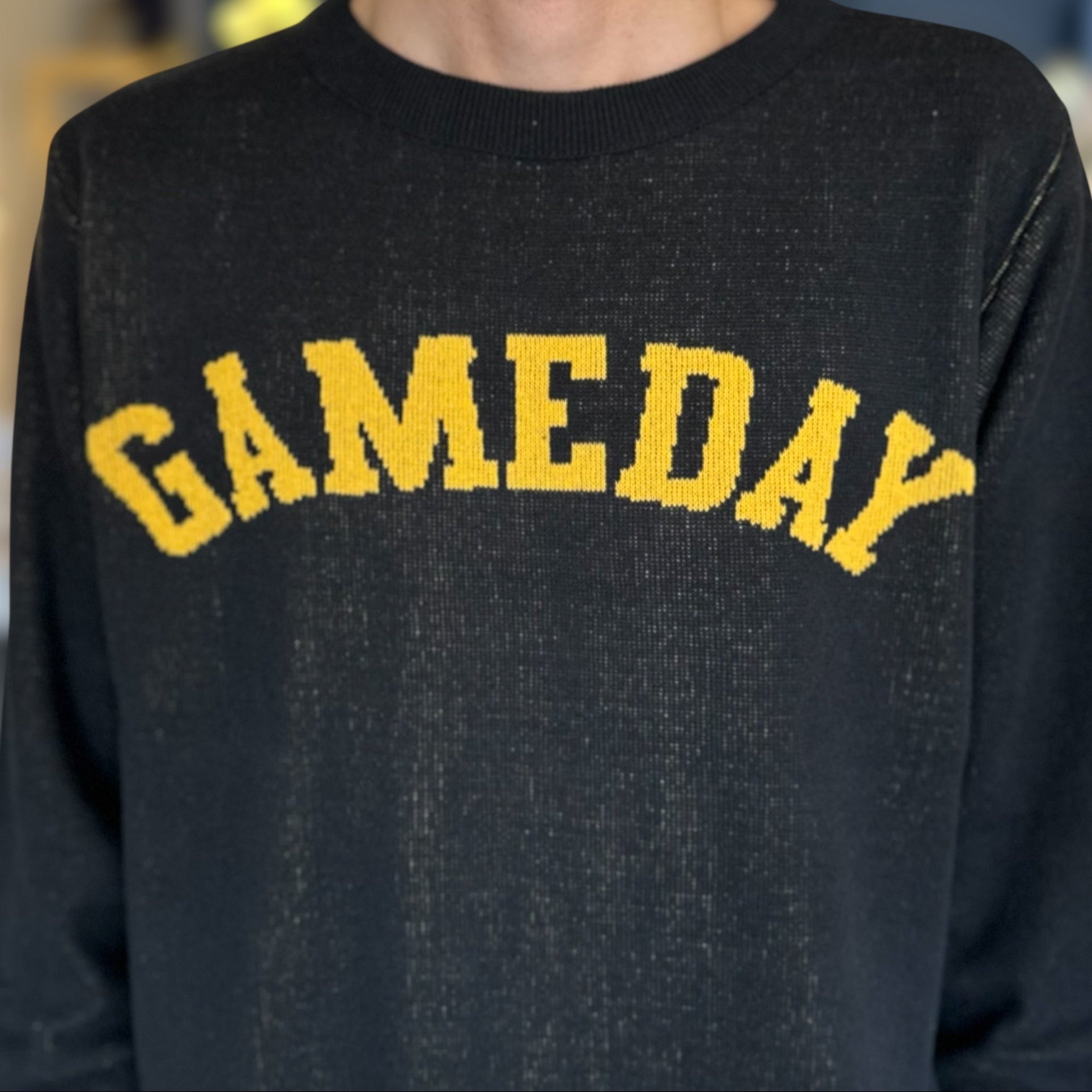 Person wearing a black sweatshirt with 'GAMEDAY' in yellow text, blurred indoor background