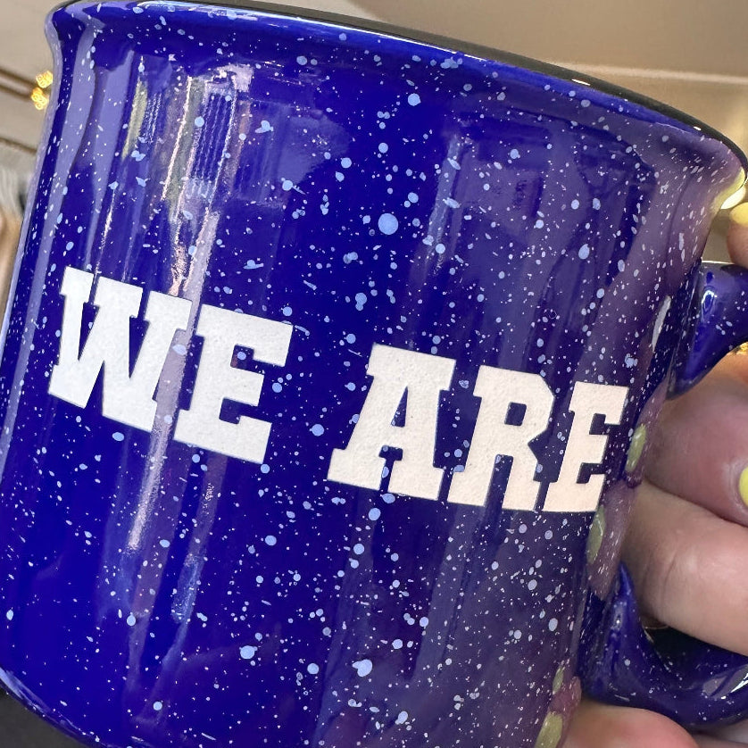 Blue mug with 'WE ARE' text held by a hand with yellow nail polish.