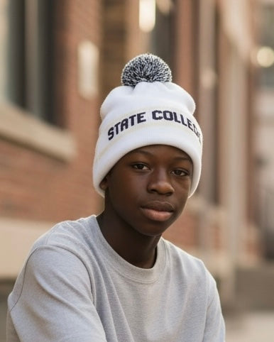 Young person wearing the white and gray STATE COLLEGE slouch pom-pom beanie while sitting on a downtown sidewalk.