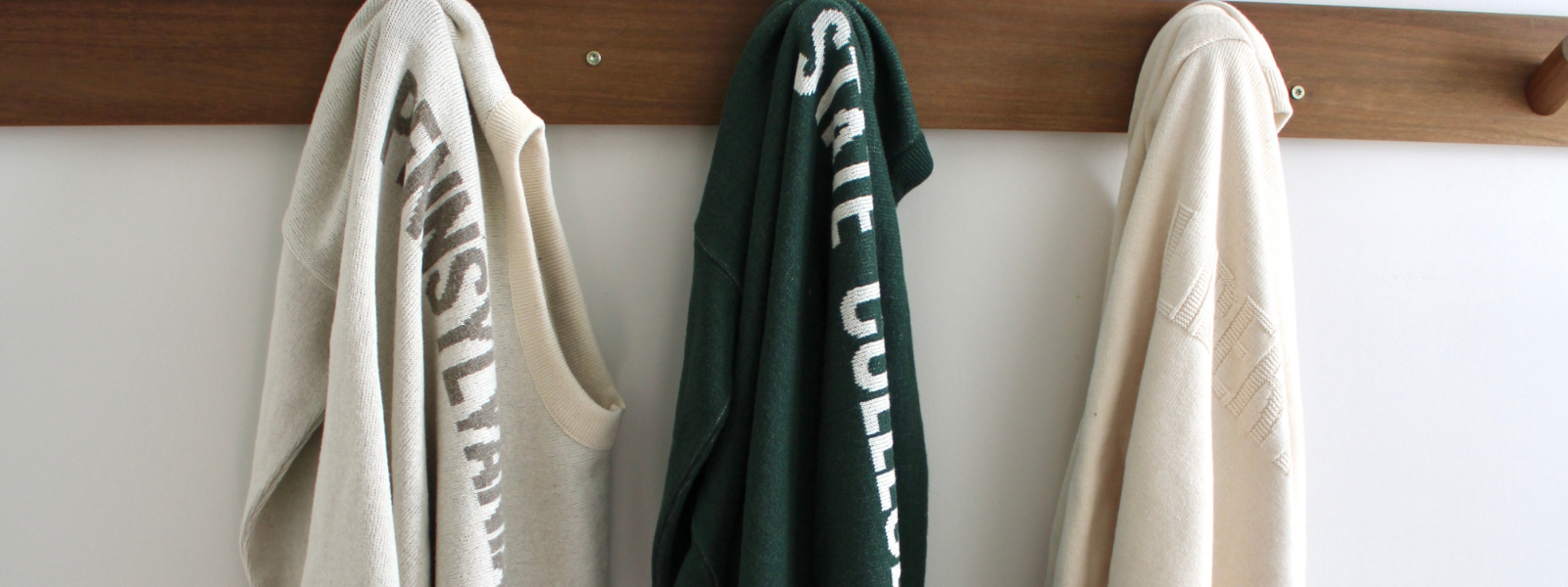 Three sweaters hanging on a wooden rack against a white wall.