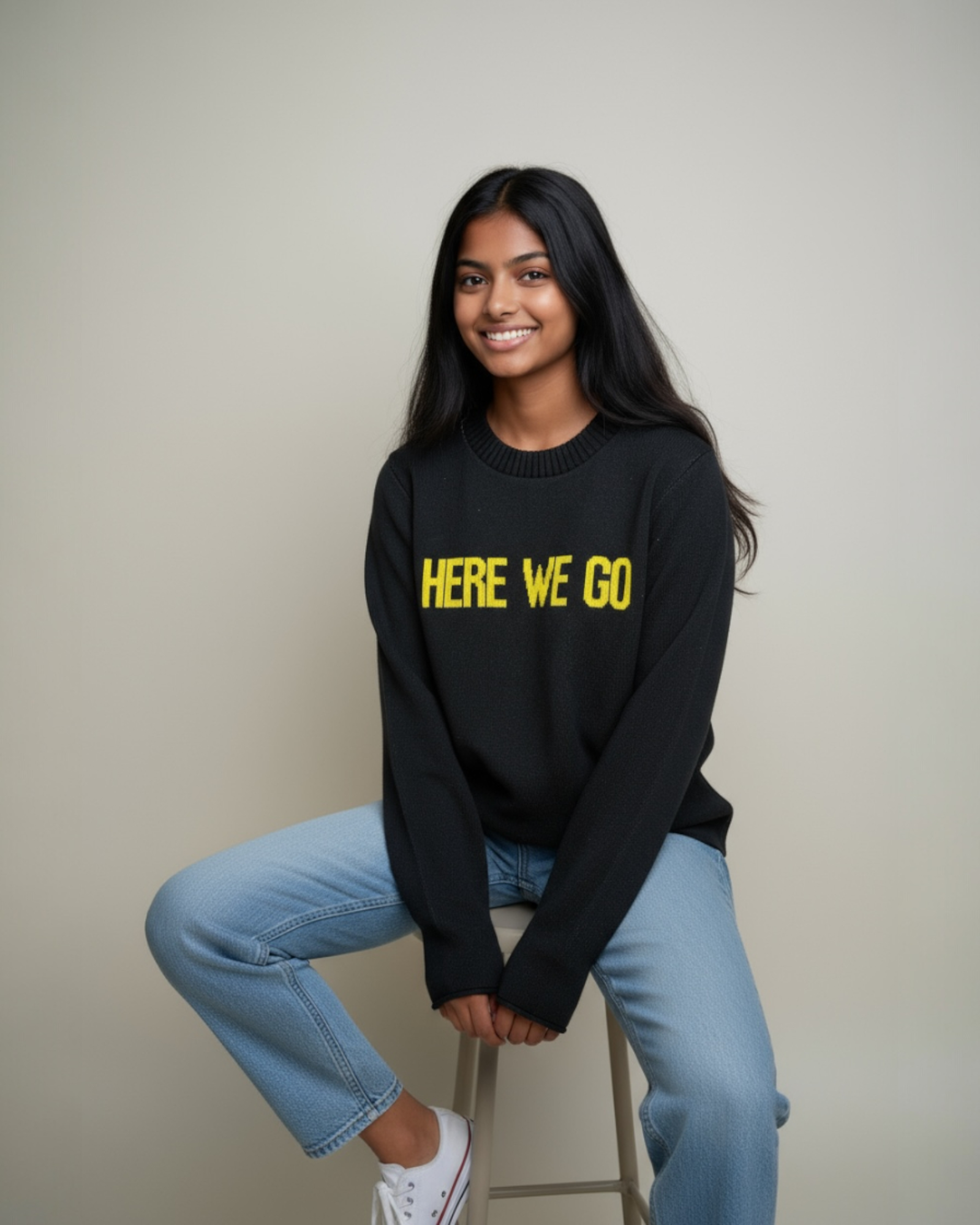Woman sitting on stool wearing black-and-gold “HERE WE GO” sweater with jeans and white sneakers. Town Pride USA.