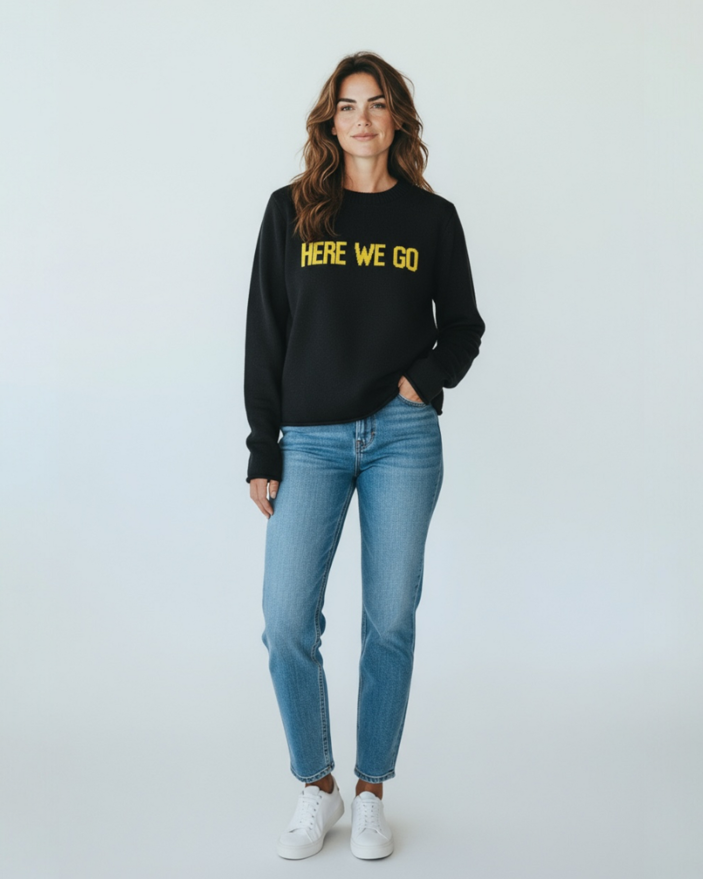Woman wearing black-and-gold “HERE WE GO” sweater by Town Pride, standing in jeans and sneakers. Made in the USA from recycled cotton.