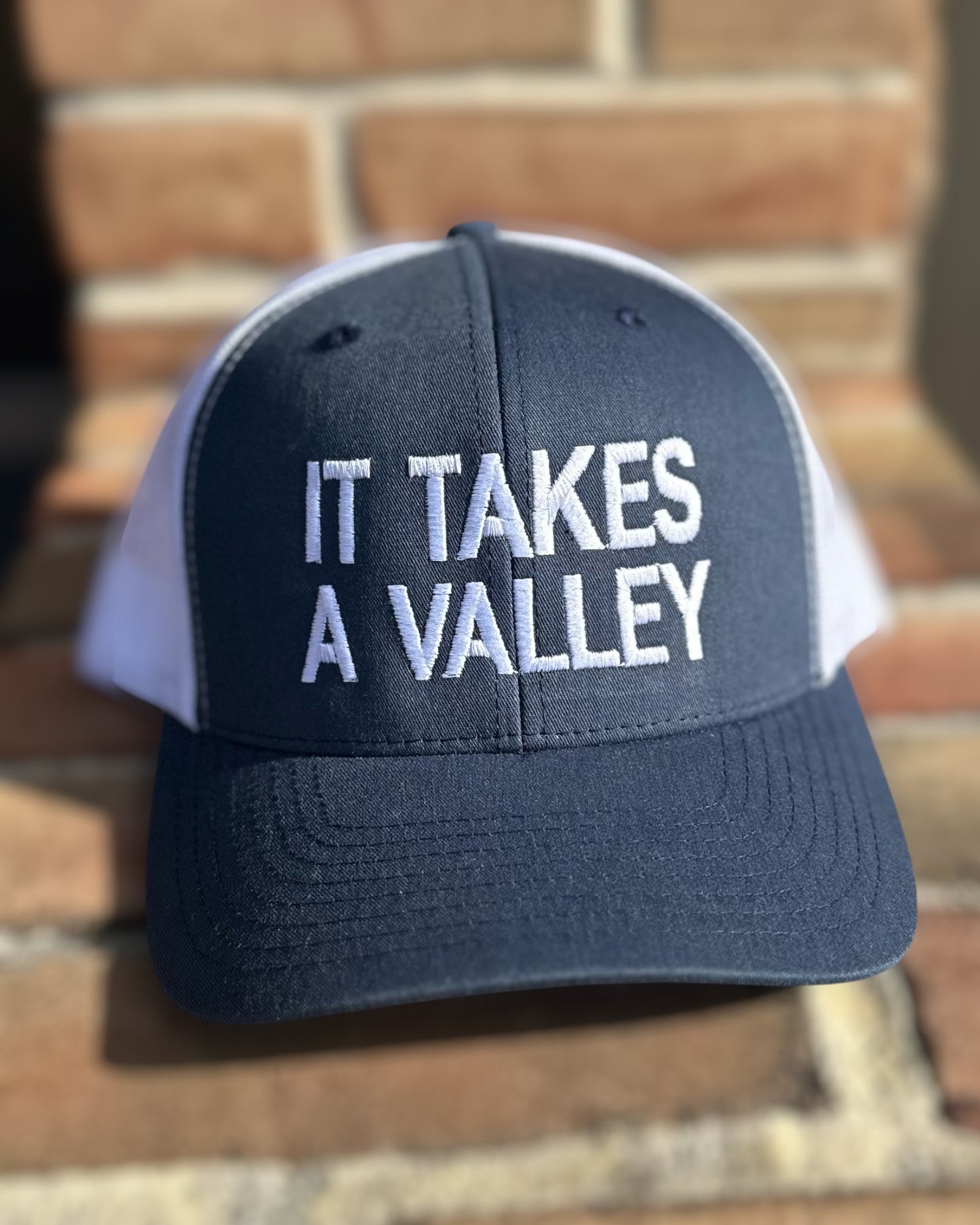 Navy and white It Takes A Valley™ trucker hat with embroidered lettering, displayed on brick background.