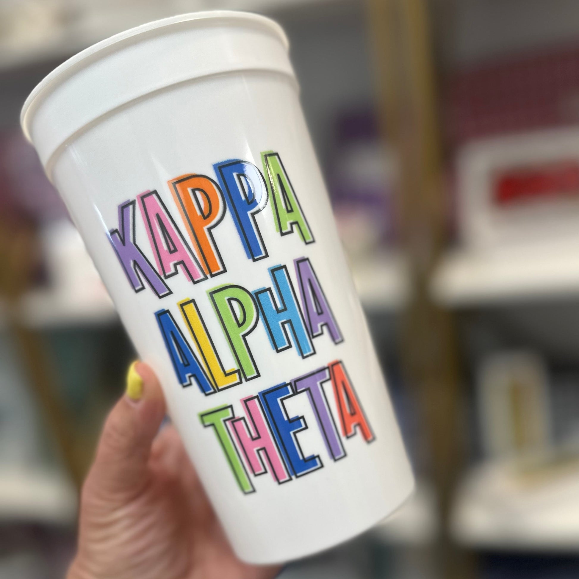 White tumbler with colorful 'Kappa Alpha Theta' text held by a person in a store setting.