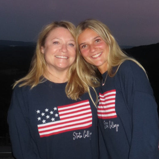 Two people wearing matching navy sweatshirts with an American flag design, standing against a dark background.