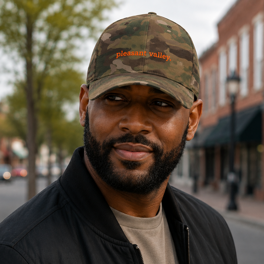 A man wearing a camouflage baseball cap with 'pleasant valley' embroidered in orange on the front, standing outdoors on a city street with brick buildings in the background.