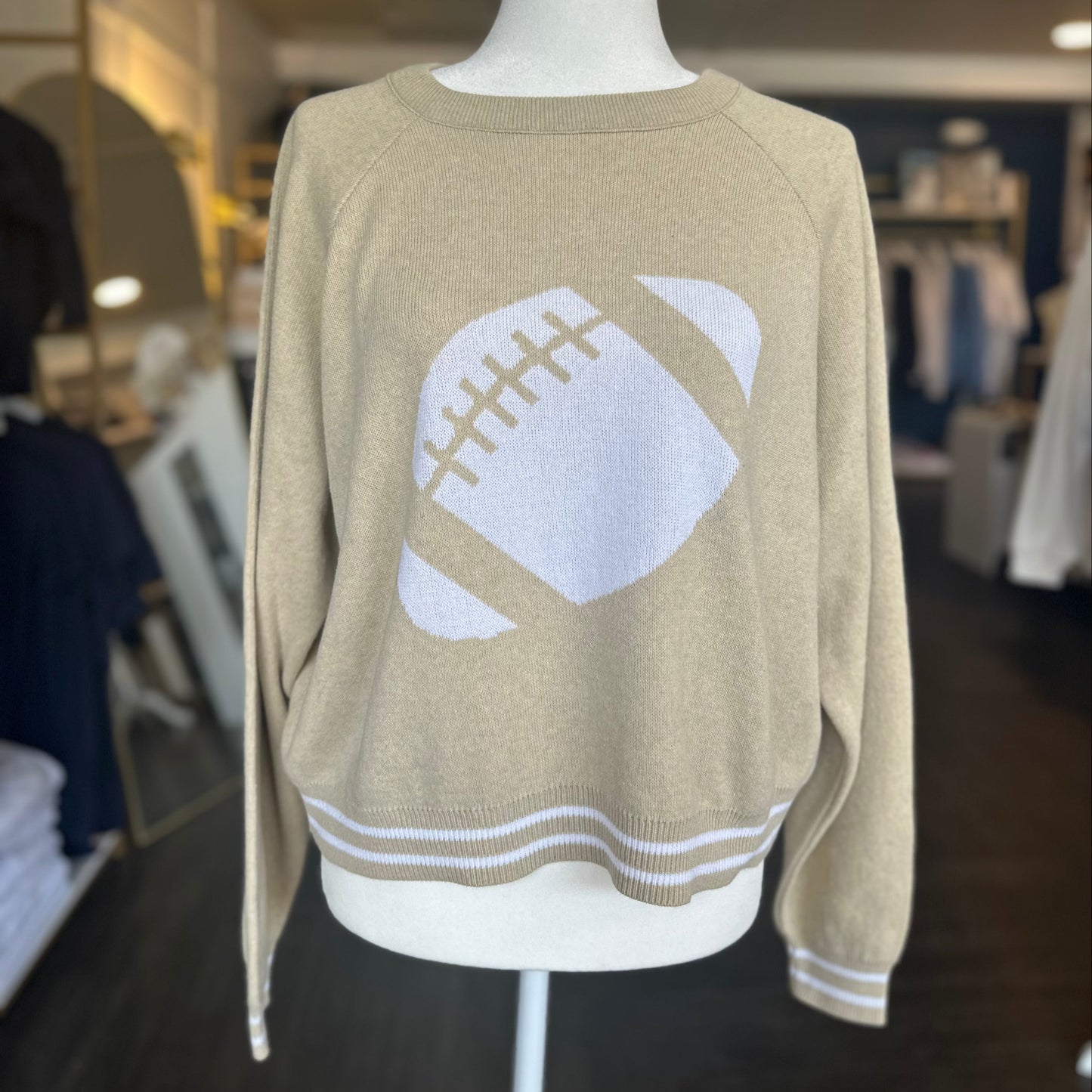 Beige sweater with a white football design on a mannequin in a store setting