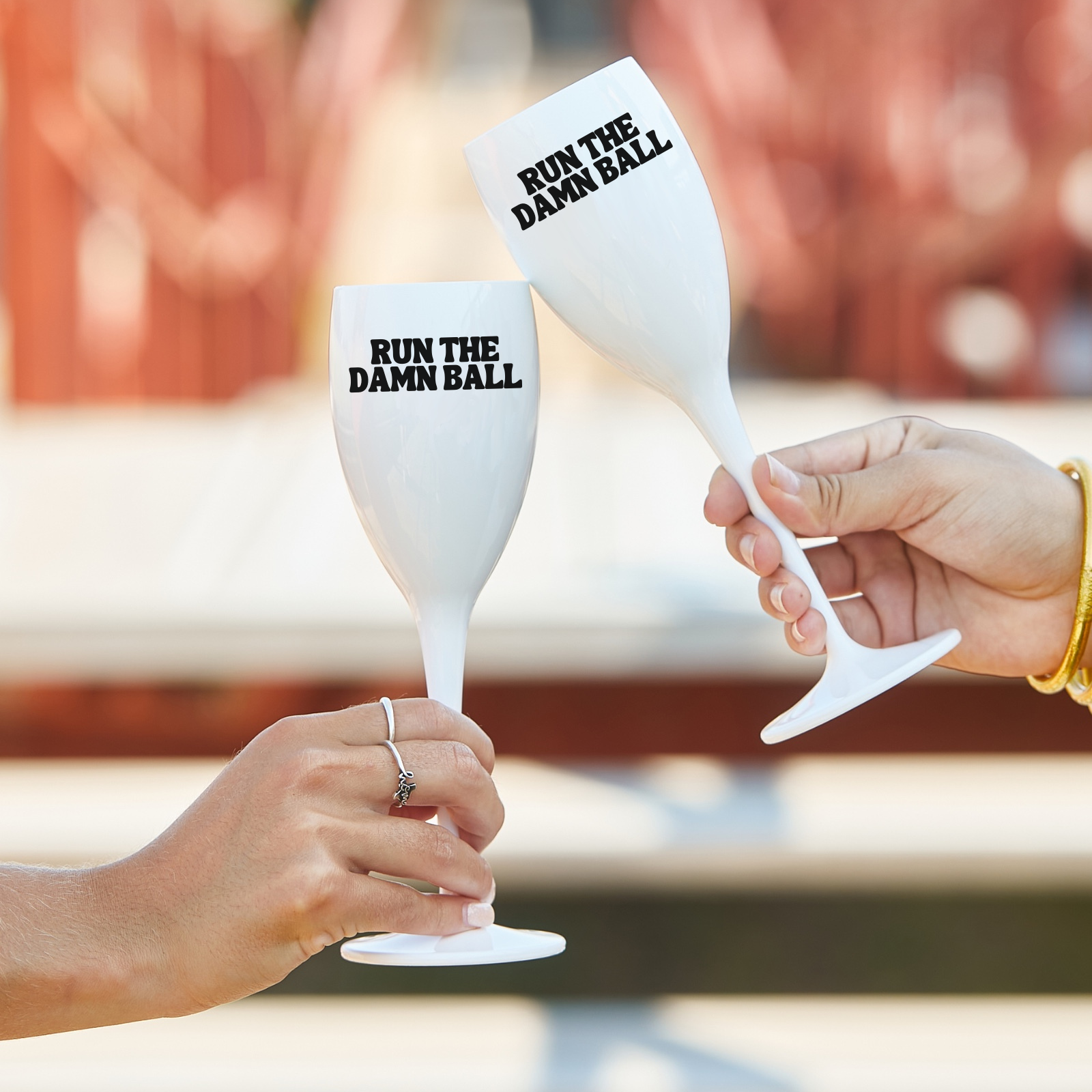 Two white wine glasses with 'RUN THE DAMN BALL' text held by hands against a blurred outdoor background.