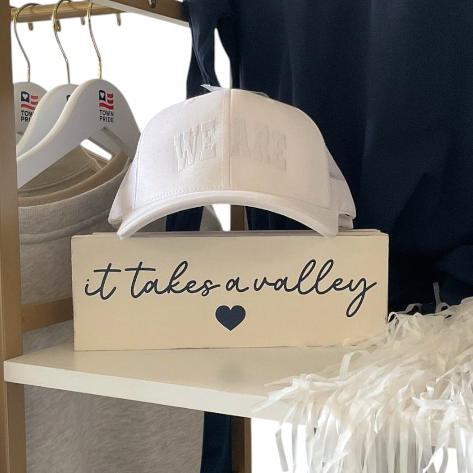 A retail display featuring navy blue and white varsity-style apparel, including a sweatshirt, t-shirt, and baseball cap with 'WE ARE' lettering, alongside a cream sign reading 'it takes a valley' and a white cap on a shelf.