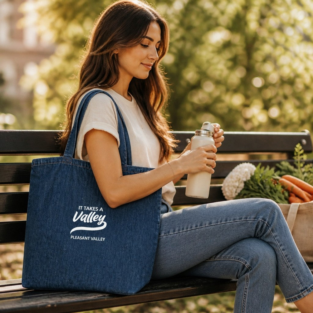 Woman sitting on a park bench holding a Town Pride State College organic denim tote with Pleasant Valley branding